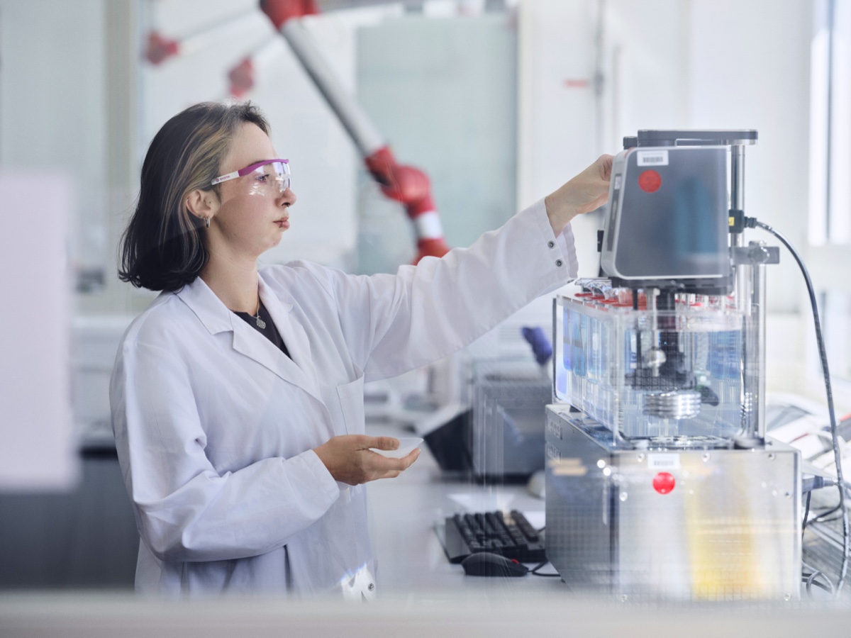 A scientist in a lab coat reaching up to adjust a laboratory instrument, a red robotic arm visible blurred in the background. Eine Wissenschaftlerin im Laborkittel justiert ein Laborgerät, im Hintergrund unscharf ein roter Roboterarm. Editorial photography / Editorialfotografie (Evonik Elements, KNSK): Robert Eikelpoth, Studio Eikelpoth, Düsseldorf.